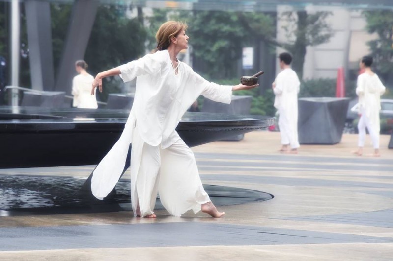 Dancer in white with blond hair extending a singing bell on outdoor beige/gray plaza in front of a granite fountain