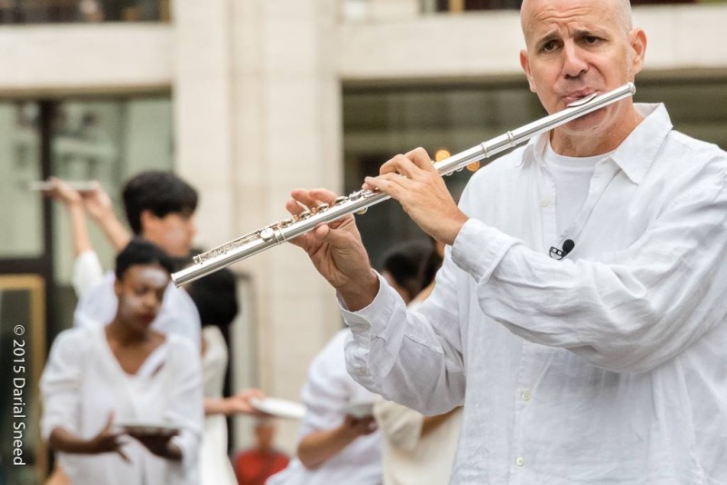Bald flautist in white with dancers in white in background in front of cream stone and glass building