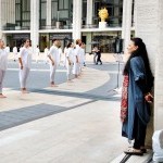 At left is a women w/ long black hair in blue/red facing dancers in white on grey/cream outdoor plaza