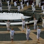 Dancers in white with one arm raised to the sky, encircling outdoor fountain on grey/beige granite plaza