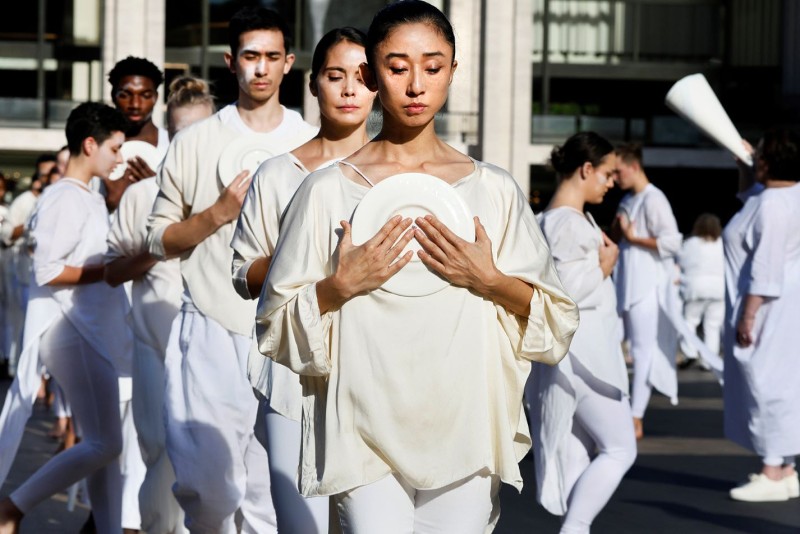 lines of dancers in white with hands clasped on chest advancing forward on outdoor plaza