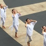 Dancers in white moving  in line formation on outside plaza, both hands vibrating by ears