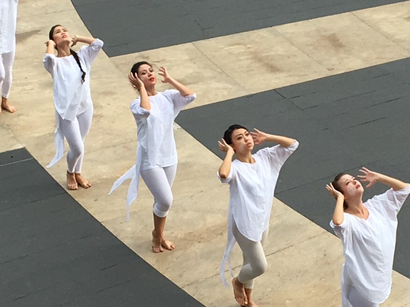 Dancers in white moving  in line formation on outside plaza, both hands vibrating by ears