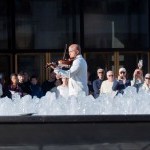 Violinist in white behind an outdoor fountain surrounded by audience