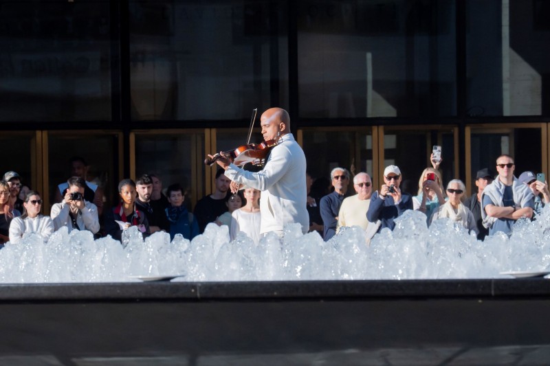 Violinist in white behind an outdoor fountain surrounded by audience