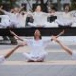 Dancers in white sitting on outdoor plaza around a circular fountain with arms upraised towards the sky