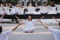 Dancers in white sitting on outdoor plaza around a circular fountain with arms upraised towards the sky
