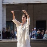 Outdoors, Dancer in white, arms upraised holding a bell in each hand