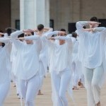 Outdoors, lines of Dancers in white with arms raised horizontally over eyes  
