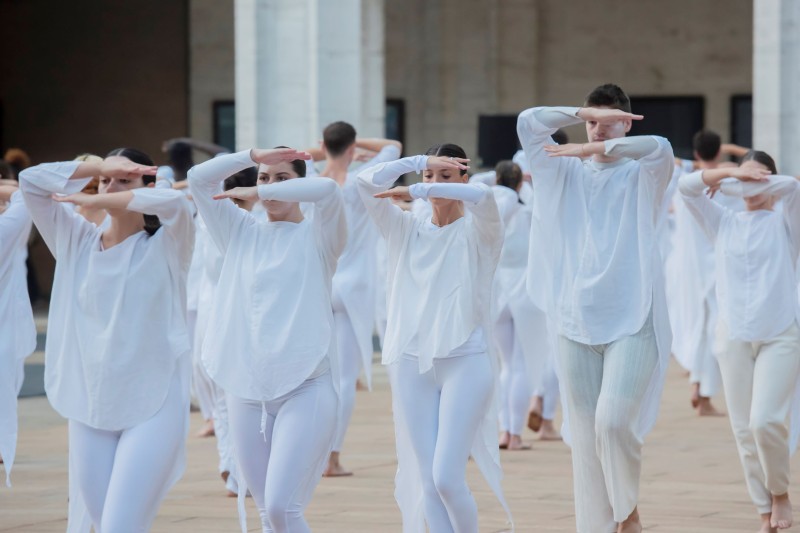 Outdoors, lines of Dancers in white with arms raised horizontally over eyes  