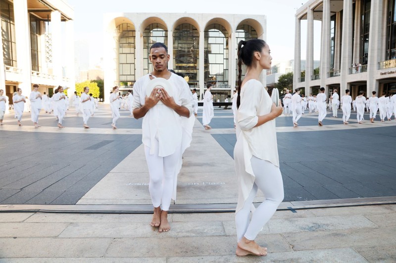 Outdoors, lines of Dancers in white clasping white ceramic plate to chest on plaza surrounded by three buildings
