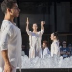Dancers in white surrounding an outdoor fountain; elevated dancer arms raised playing a singing bowl