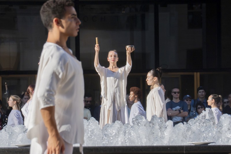 Dancers in white surrounding an outdoor fountain; elevated dancer arms raised playing a singing bowl