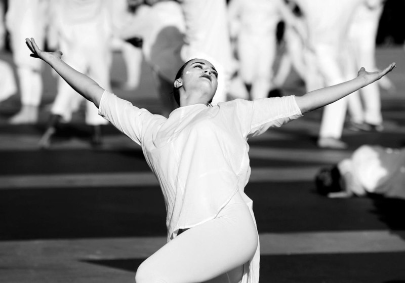 Dancer in white leaning backwards with arms raised, surrounded on outdoor plaza by dancers in white