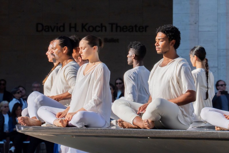 Dancers in white seated cross-legged around the rim of an outdoor black marble fountain  