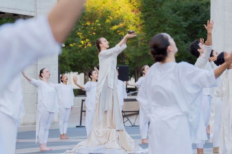 Dancers in white clasping a white ceramic plate to their chest, encircling an outdoor plaza