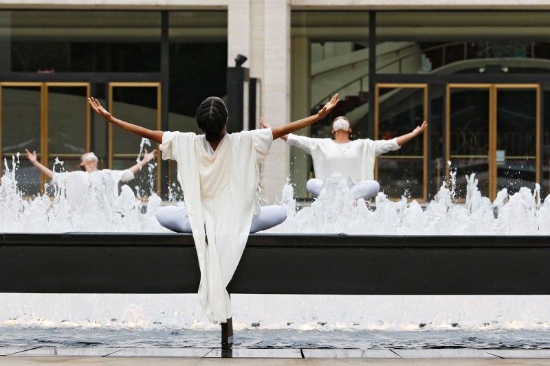 2 Dancers in white sitting on a black granite fountain with arms raised outwards towards the sky