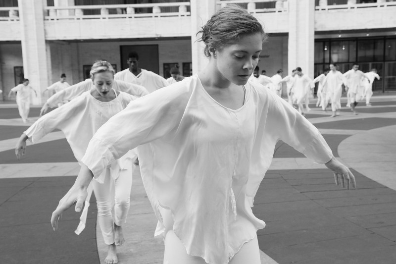 (Center) Dancer in white, arms upraised with a bell, surrounded by dancers encircling a fountains 