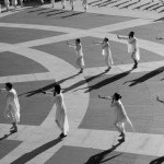 Dancers in white moving  in line formation on outside plaza, hand extended to cover mouth