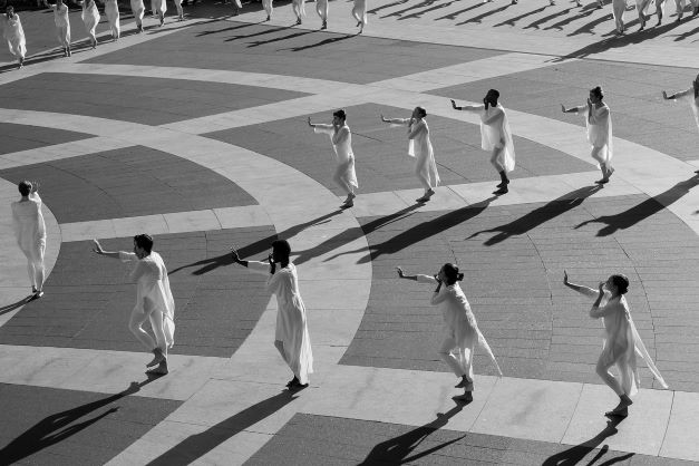 Dancers in white moving  in line formation on outside plaza, hand extended to cover mouth