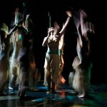 7 women in moss green costumes holding bamboo sticks reaching towards with sky 