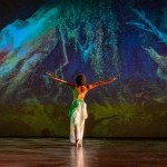 female dancer in white facing green/blue projection of roots of trees