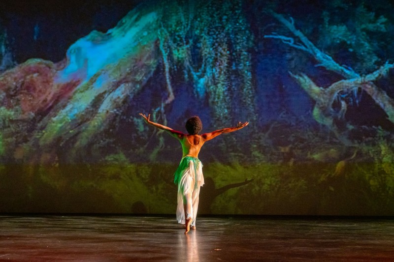 female dancer in white facing green/blue projection of roots of trees