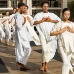 lines of dancers in white with hands clasped on chest advancing forward on plaza