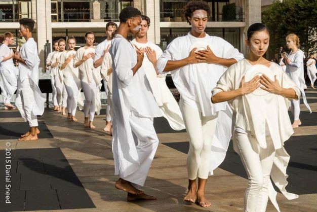 Lines of dancers in white, arms clapsing a plate against chest, outdoor grey/beige plaza