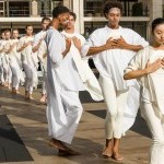 lines of dancers in white with hands clasped on chest advancing forward on outdoor plaza