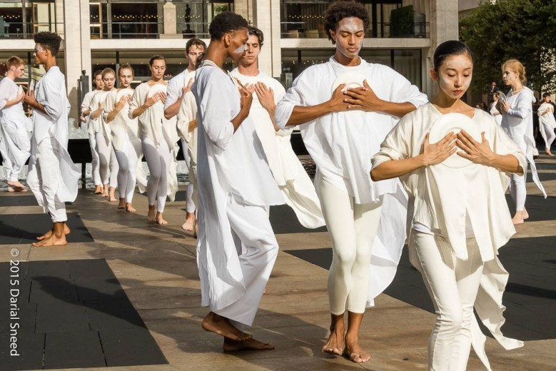 lines of dancers in white with hands clasped on chest advancing forward on outdoor plaza