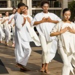 lines of dancers in white with hands clasped on chest advancing forward on plaza