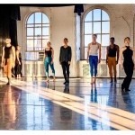Multiple dancers facing forward in front of two windows with light beam on the floor 