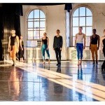 2 rows of dancers facing upstage with windows in background and a diagonal light beam on the floor