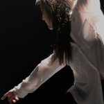 Dancer in white long sleeve shirt dancing onstage in spotlight with black background.