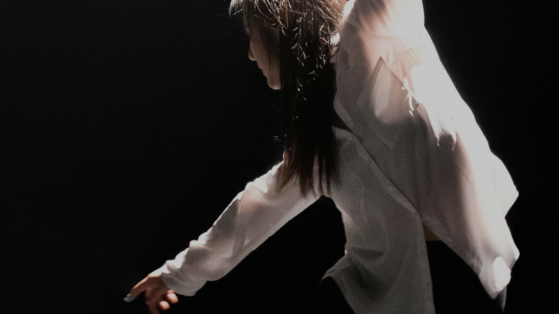Dancer in white long sleeve shirt dancing onstage in spotlight with black background.