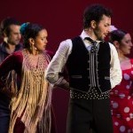 A group of flamenco dancers in full costume. Each is holding their arms behind their back and looking over their left shoulder.