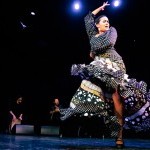 A flamenco dancer in a black, white and flower patterned dress raises her right arm and looks over her left shoulder.