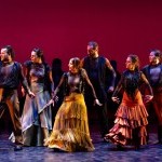 Six flamenco dancers in earth tone colored costumes stand in a group looking to the left.
