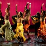 Dancers in brightly colored costumes face the left with their arms up.