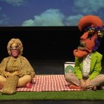 Yali Romagoza & Autumn Newcomb, sit on a red and white checkered blanket over fake grass with a projection of clouds behind them