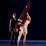 three dancers wearing black, red, and beige. one dancer is lifted upside down.