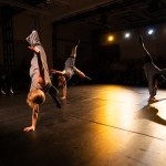 three dancers inverted in handstands, surrounded by audience spectators