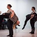 Four dancers are in front of an audience. Two dancers are being lifted and carried in two dancer's arms. 