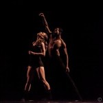 Image of 2 dancers against a dark background. One dancer's hand is above the other's head, which is looking up towards the hand.