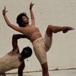 Two contemporary dancers pose against a wall, one balancing with a raised leg while the other crouches beneath in support.