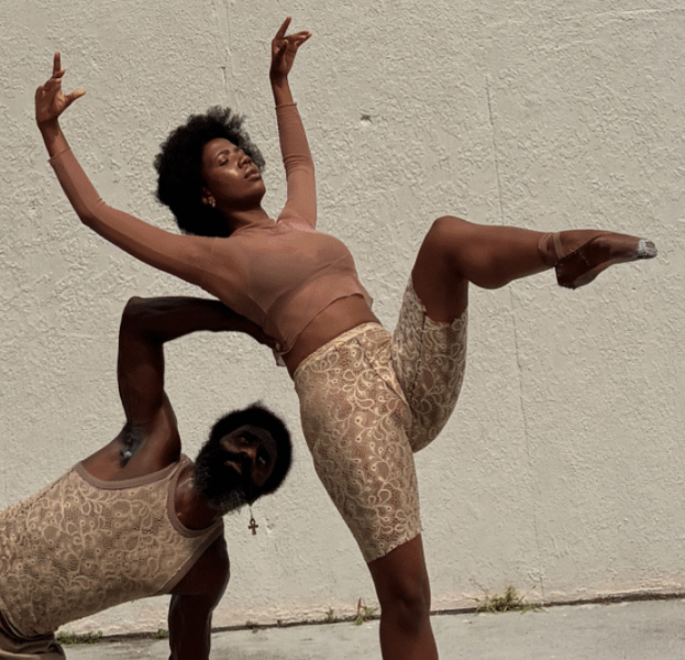 Two contemporary dancers pose against a wall, one balancing with a raised leg while the other crouches beneath in support.