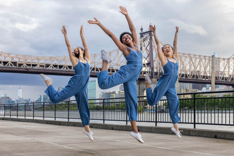 3 female dancers in blue jumpsuits, jumping in air arms overhead, Queensborough bridge in background