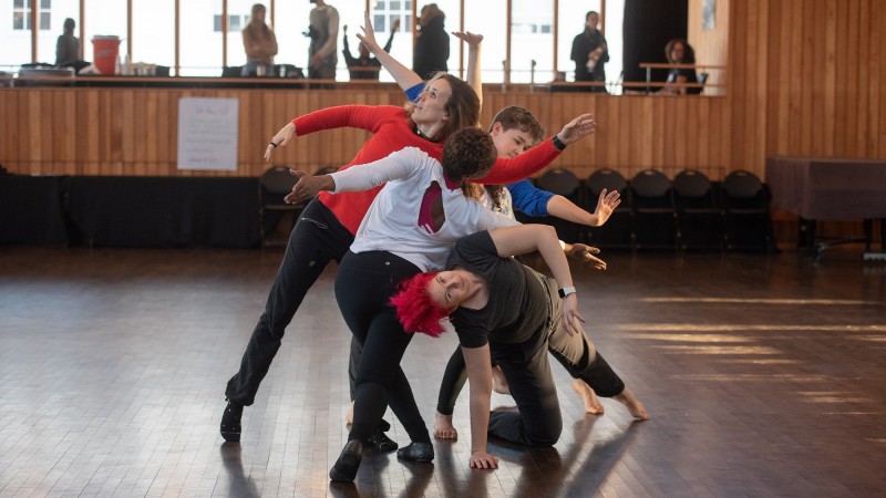 A group of four dancers are clumped together and create an abstract shape with their bodies in a wood-paneled dance studio.