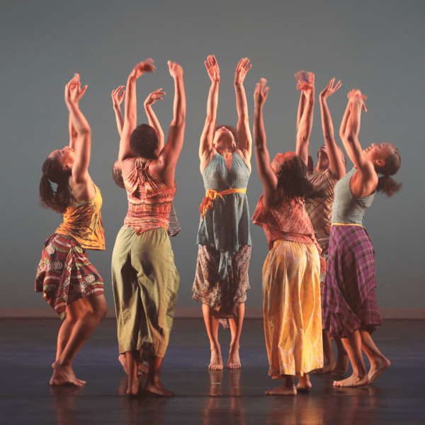 Seven female presenting dancers in a circle, looking towards the ceiling, with their arms raised above their heads.